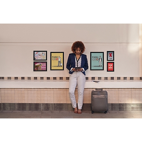 Une femme aux cheveux bouclés s'appuie contre un mur avec des affiches et regarde son smartphone, une valise à côté d'elle.