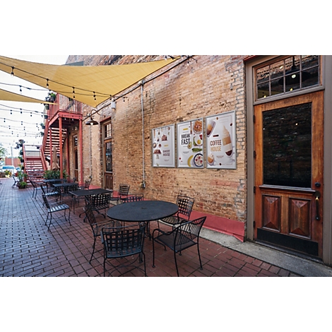 Espace extérieur d'un café avec tables, chaises et menus affichés sur un mur en briques sous une voile d'ombrage.
