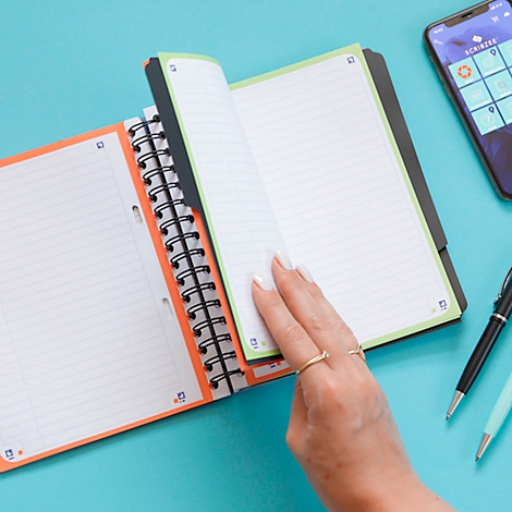Gros plan : une personne feuillette un cahier avec des accents verts et oranges. Un téléphone et des stylos sont sur la table.