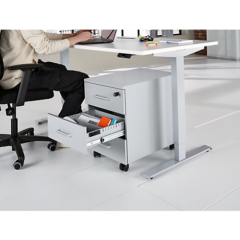 Une personne est assise à un bureau réglable en hauteur et travaille sur un ordinateur portable. Le tiroir du caisson mobile est ouvert.