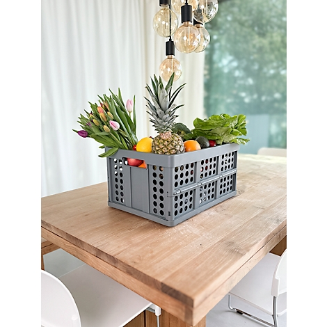 Caja gris llena de frutas y flores sobre una mesa de madera, con una lámpara colgando encima. Dos sillas están preparadas.
