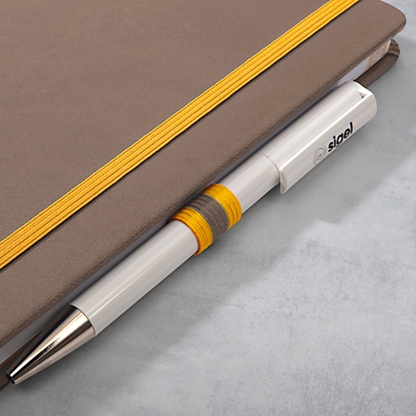 Cahier gris avec des bandes élastiques jaunes. Stylo blanc avec impression "sigel" dans le livre.