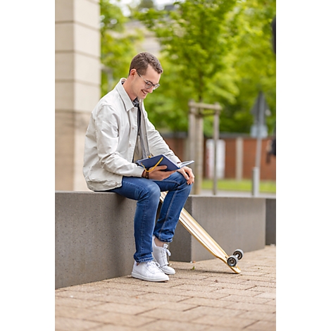 Un homme est assis avec un carnet et un longboard. Il sourit, porte des lunettes, un jean et une veste légère.