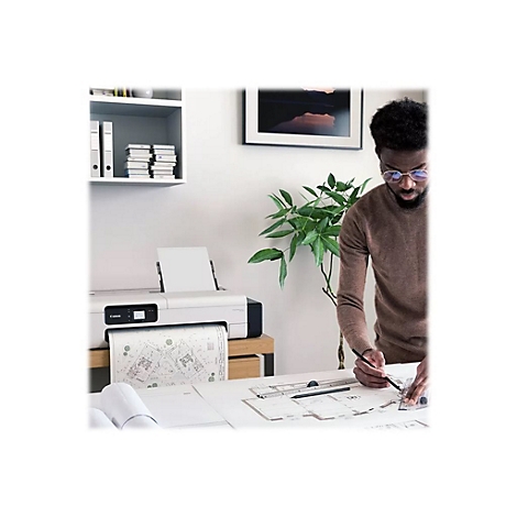 Ein Mann mit Brille zeichnet mit Stift an einem Tisch, auf dem eine technische Zeichnung liegt. Im Hintergrund ein Drucker, eine Pflanze und ein Bild.
