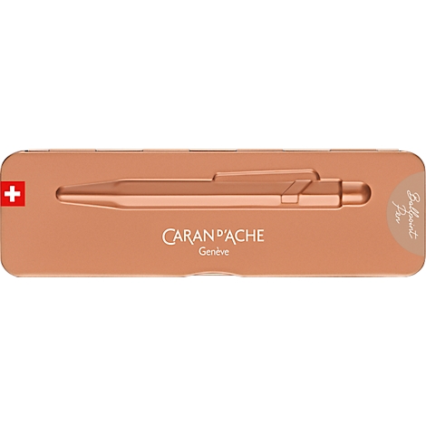 Kupferfarbener Stift in einer länglichen, rosafarbenen Schachtel. Schweizer Flagge. Schriftzug: Caran d'Ache Genève, Ballpoint Pen.
