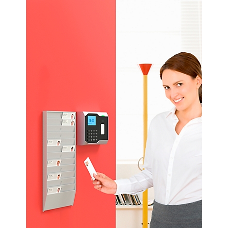 A woman holds a card in front of a time clock on a red wall. The card shows a photo and names.