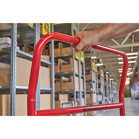 Carretilla roja en un almacén. Un brazo sostiene el asa. Estanterías con cajas al fondo.