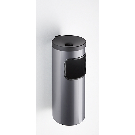 Poubelle cylindrique grise avec couvercle et ouverture noirs. Un champ sombre avec des coutures décoratives. Fixé à un mur blanc.