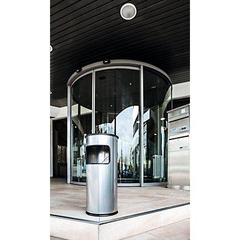 Une poubelle cylindrique en acier inoxydable se trouve devant une porte tambour en verre à une entrée moderne.
