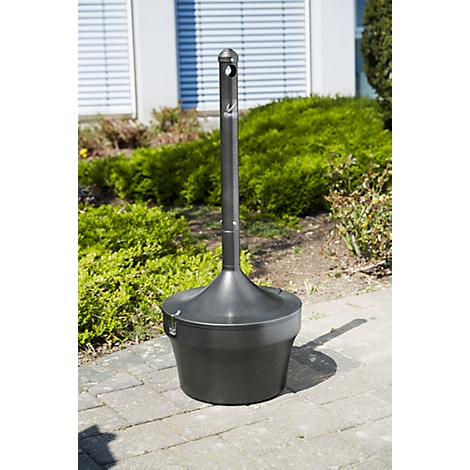Cendrier haut gris foncé avec support à l'extérieur. Un logo de marque est visible sur le support. Il se trouve sur un chemin pavé, avec des plantes vertes en arrière-plan.