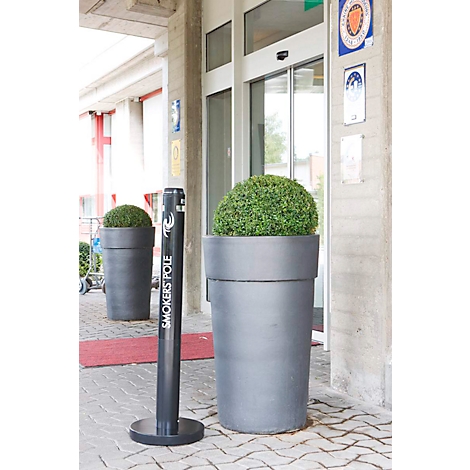 Des jardinières gris foncé avec des buissons verts flanquent une porte vitrée. Un "Smokers Pole" noir se trouve devant.