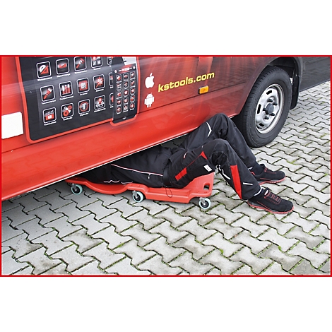 Un mécanicien est couché sous un véhicule rouge sur un chariot, les jambes sortant. Sur le véhicule, il est écrit ks-tools.com.