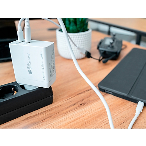 Chargeur blanc branché dans une prise de courant noire. À côté, une tablette et une plante en pot.