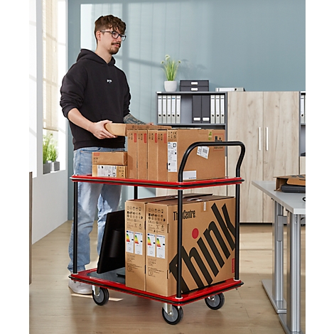 Un homme avec des lunettes charge un chariot de transport rouge avec des colis. Des boîtes et un moniteur sont sur le chariot.