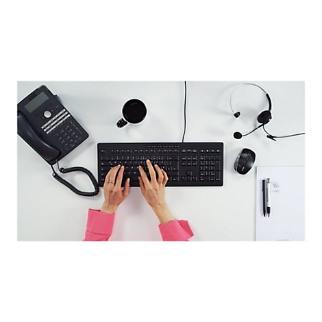 Person tippt auf einer schwarzen Tastatur. Auf dem Schreibtisch: Telefon, Headset, Maus, Becher, Stifte, Papier.