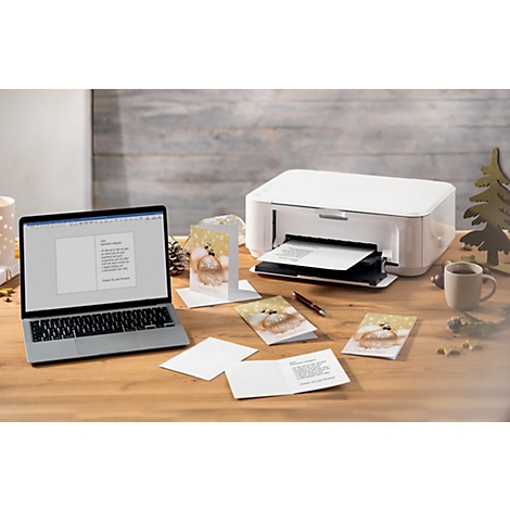 Laptop, printer, and Christmas cards on a wooden table. The screen displays text being printed. Several finished cards are beside it.