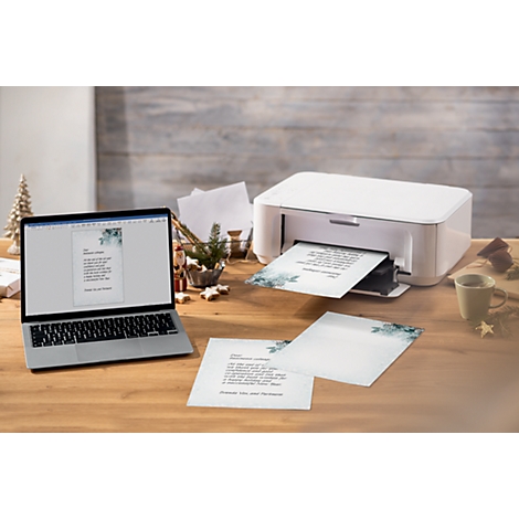 A laptop shows a printed document, next to it a white printer is printing more sheets. Finished sheets are lying on the table.