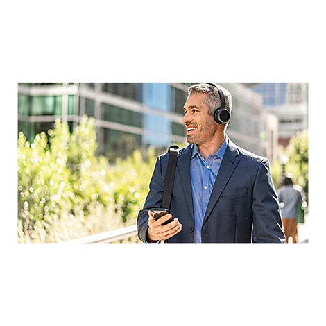 Un homme souriant portant un casque, portant une mallette, tenant un smartphone, des bâtiments et des espaces verts en arrière-plan.