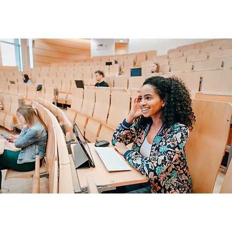 Une femme souriante est assise dans un amphithéâtre. Elle a les cheveux bouclés et porte une veste à motifs floraux. Il y a une tablette, un clavier et une souris sur le bureau.