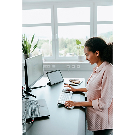 Une femme est assise à un bureau. Elle tape sur un clavier, à côté d'un moniteur et d'une tablette. Derrière elle, il y a une fenêtre avec des arbres.