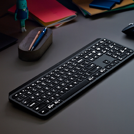 Clavier noir avec touches éclairées sur un bureau gris. A côté : stylos, cahiers.