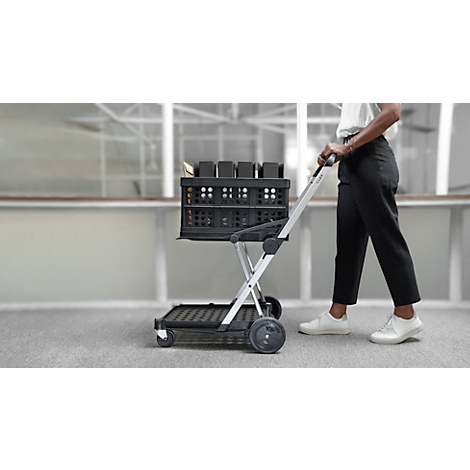 Person pulling a black shopping cart. The cart is filled with boxes. Person is wearing a white blouse, black trousers, and white shoes.