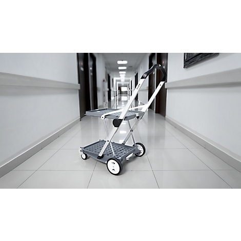 A gray, foldable shopping cart stands in a bright hallway. White wheels and handle.