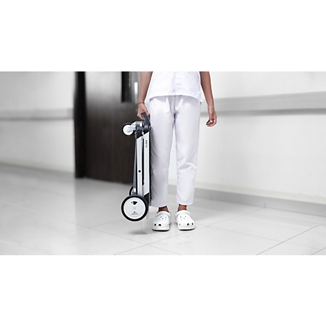A person in white holding a folded wheelchair. They are standing in a white hallway.