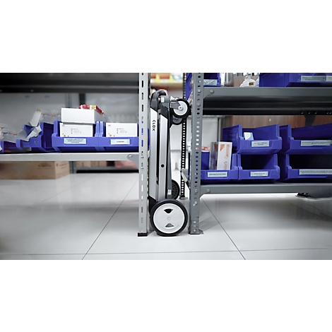 A silver transport trolley stands between warehouse shelves with blue bins, wheels and chain.