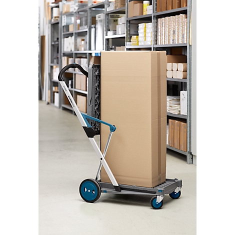 Ein grauer Transportwagen mit blaugrünen Rädern und Griffen transportiert einen grossen Karton in einem Lagerraum.