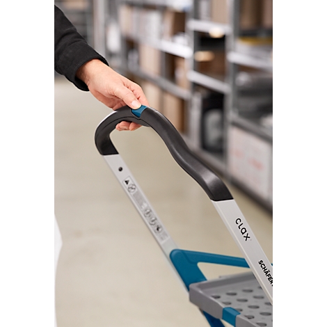 Nahaufnahme einer Hand, die den Griff eines Einkaufswagens hält. Der Griff ist schwarz und blau, dahinter Regale.