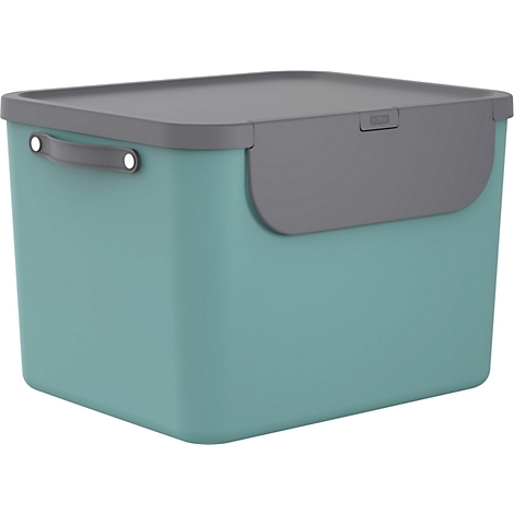 Une femme jette du carton dans une poubelle verte et noire dans une cuisine. Elle porte un jean et un haut.