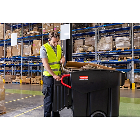 Un homme dans un entrepôt avec un gilet de sécurité et un chariot à ordures noir. Il y met du carton.