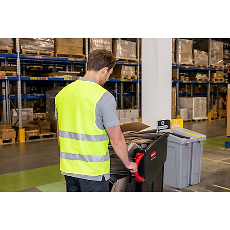 Un homme en gilet de sécurité pousse un chariot de déchets noir dans un entrepôt. Des étagères avec des marchandises sont visibles en arrière-plan.