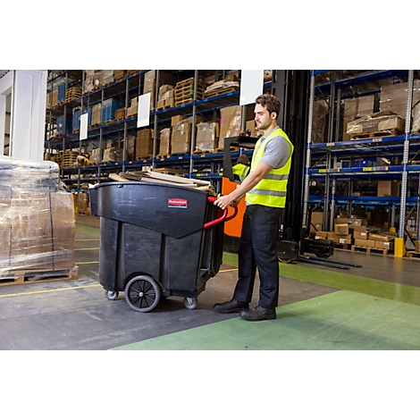 Un homme pousse un chariot à ordures noir dans un entrepôt. Il porte un gilet de sécurité.