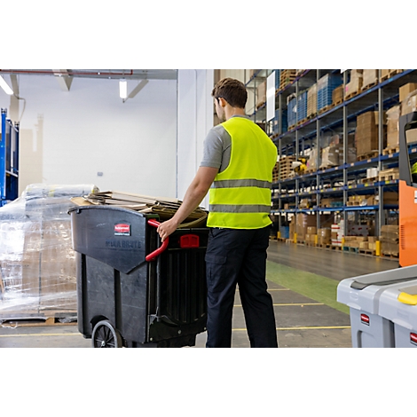 Un homme en gilet de sécurité pousse un chariot à ordures noir dans un entrepôt. Il y a du carton à l'intérieur.