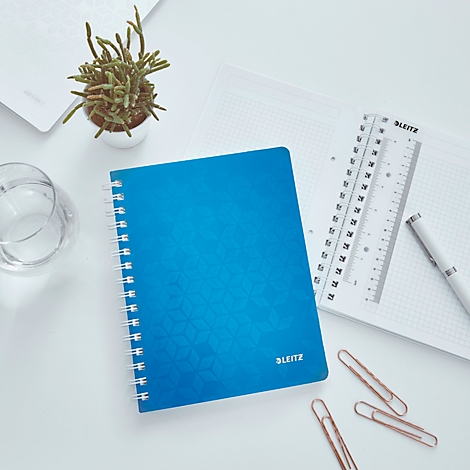 Blaues Notizbuch auf weißem Tisch. Daneben Lineal, Stift, Büroklammern und Topfpflanze. Auf dem Cover: 'LEITZ'.