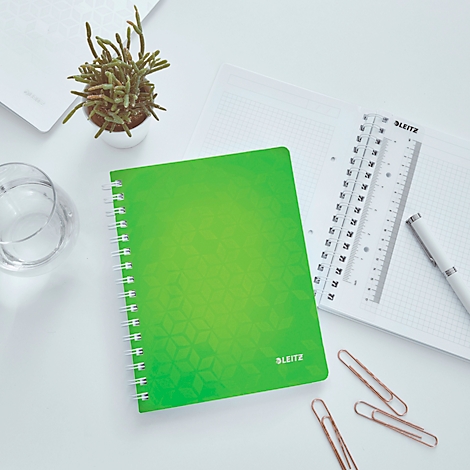 Grünes Notizbuch mit Spirale, daneben Lineal, Stift und Büroklammern, auf weißem Tisch.