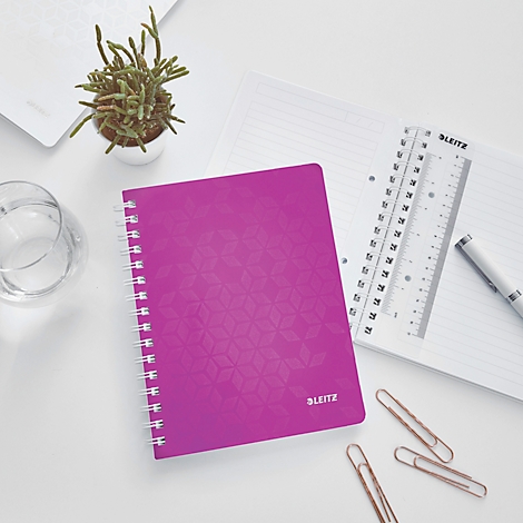 Pinkes Notizbuch mit Spiralbindung auf einem Schreibtisch. Daneben: Pflanzen, Stift, Lineal, Papier, Büroklammern.