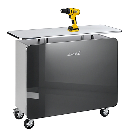 Un meuble gris à roulettes avec des bords argentés et un plateau en verre, sur lequel se trouve une perceuse jaune. Le mot "cool" est gravé sur le devant.