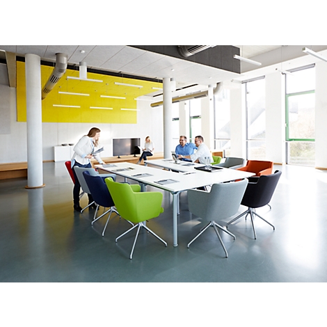 A conference room with people. A woman stands at the table, others are sitting and working. Colorful chairs.