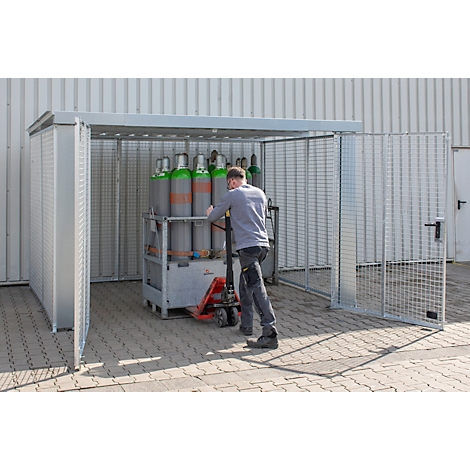 Un homme pousse des bouteilles de gaz sur un transpalette dans une enceinte de sécurité métallique. Enceinte avec porte grillagée ouverte.