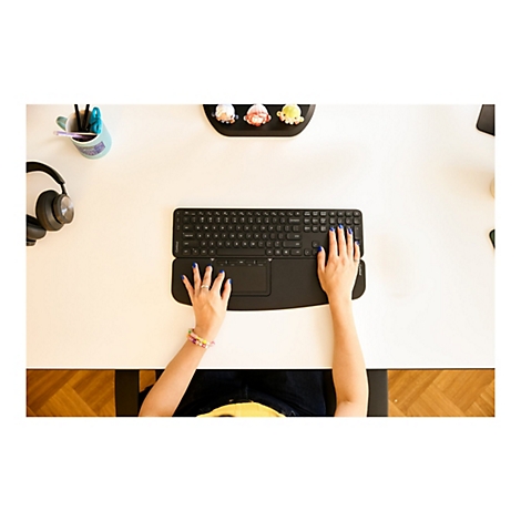 Ansicht von oben auf eine Person, die eine schwarze ergonomische Tastatur mit integriertem Trackpad benutzt, blaue Fingernägel. Auf dem Tisch stehen Stifte, eine Tasse und ein Kopfhörer.