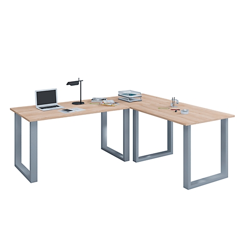 L-shaped desk with a light brown table top and gray metal legs, on which there is a laptop, a lamp, and books.