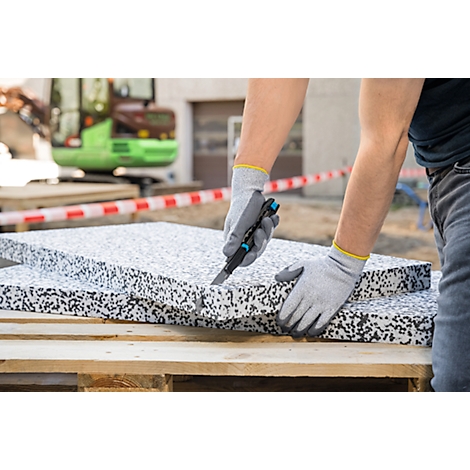 Personne coupe l'isolant avec un couteau. Gants gris avec poignets jaunes, palettes en arrière-plan.