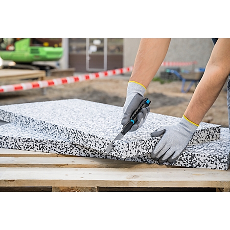 Mains gantées coupant des panneaux isolants avec un cutter. L'arrière-plan montre un chantier.
