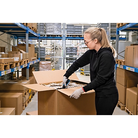 Une femme en tenue de travail ouvre avec concentration un carton d'expédition dans un entrepôt entre des rayonnages à palettes.