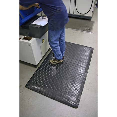 A man stands on a black, patterned workplace mat. In the background, a machine and gray floor.