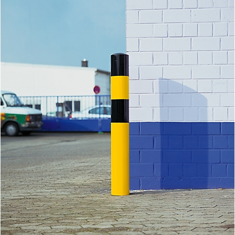 Yellow and black bollard in front of white and blue brickwork. Vehicles and a gate in the background.