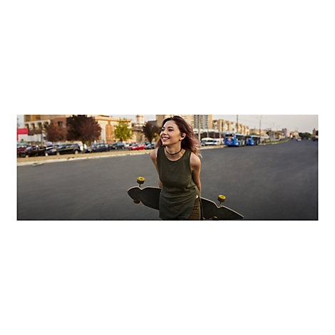 Junge Frau mit Longboard lÃ¤chelt auf der StraÃŸe. Sie trÃ¤gt KopfhÃ¶rer, ein grÃ¼nes Top und eine Halskette.